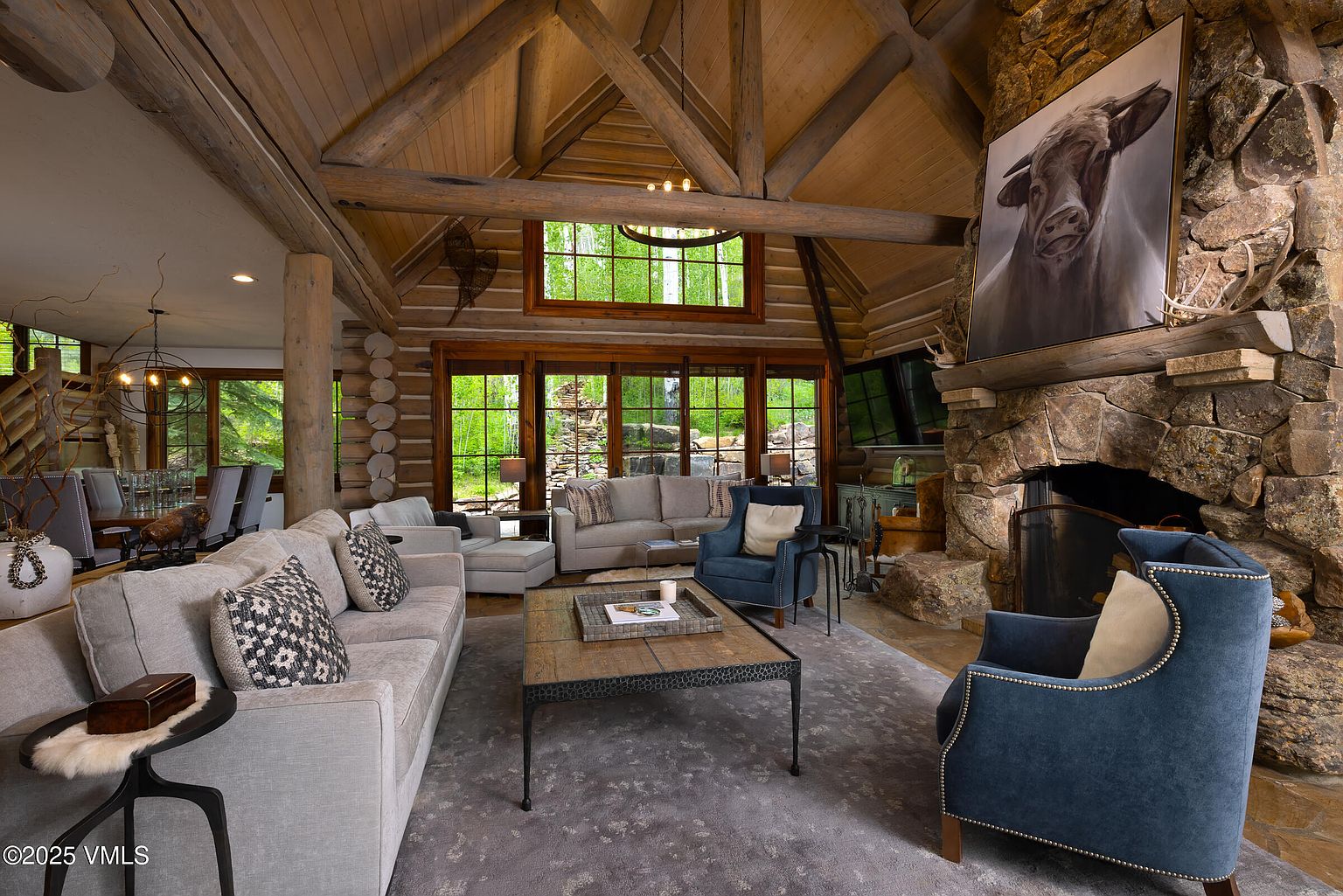This is a spacious living room featuring a high vaulted ceiling with exposed wooden beams and log walls, creating a rustic and inviting atmosphere. The room is furnished with comfortable sofas, armchairs, and a large coffee table, all arranged around a stone fireplace with a large painting above it. Large windows offer views of the surrounding greenery, enhancing the connection with nature.