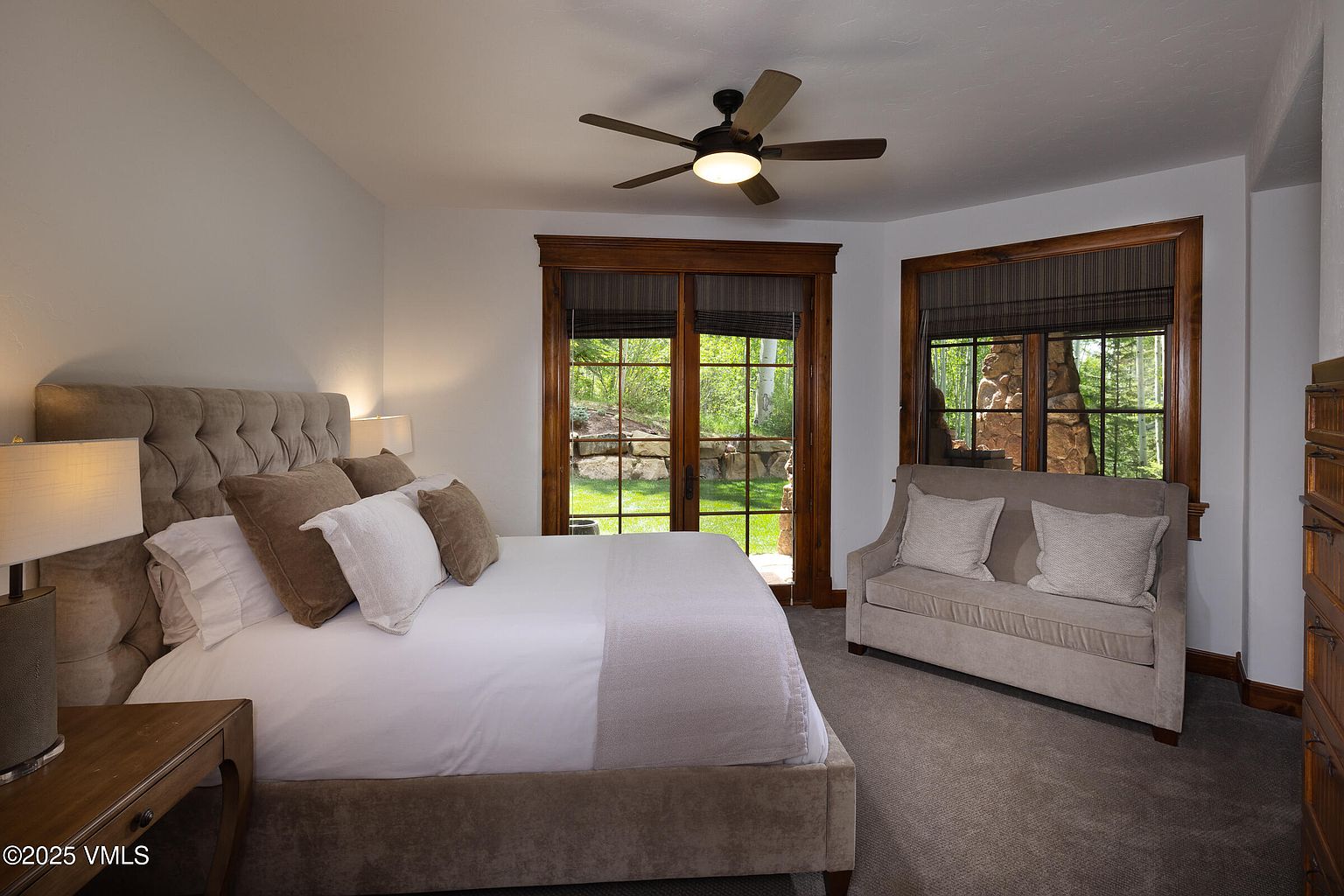 This is a primary bedroom featuring a large, tufted headboard bed with neutral-toned bedding and pillows. The room has a ceiling fan and natural light coming through a set of French doors and a window, both framed in dark wood. A small settee sits near the window, and the overall impression is one of comfort and understated elegance.