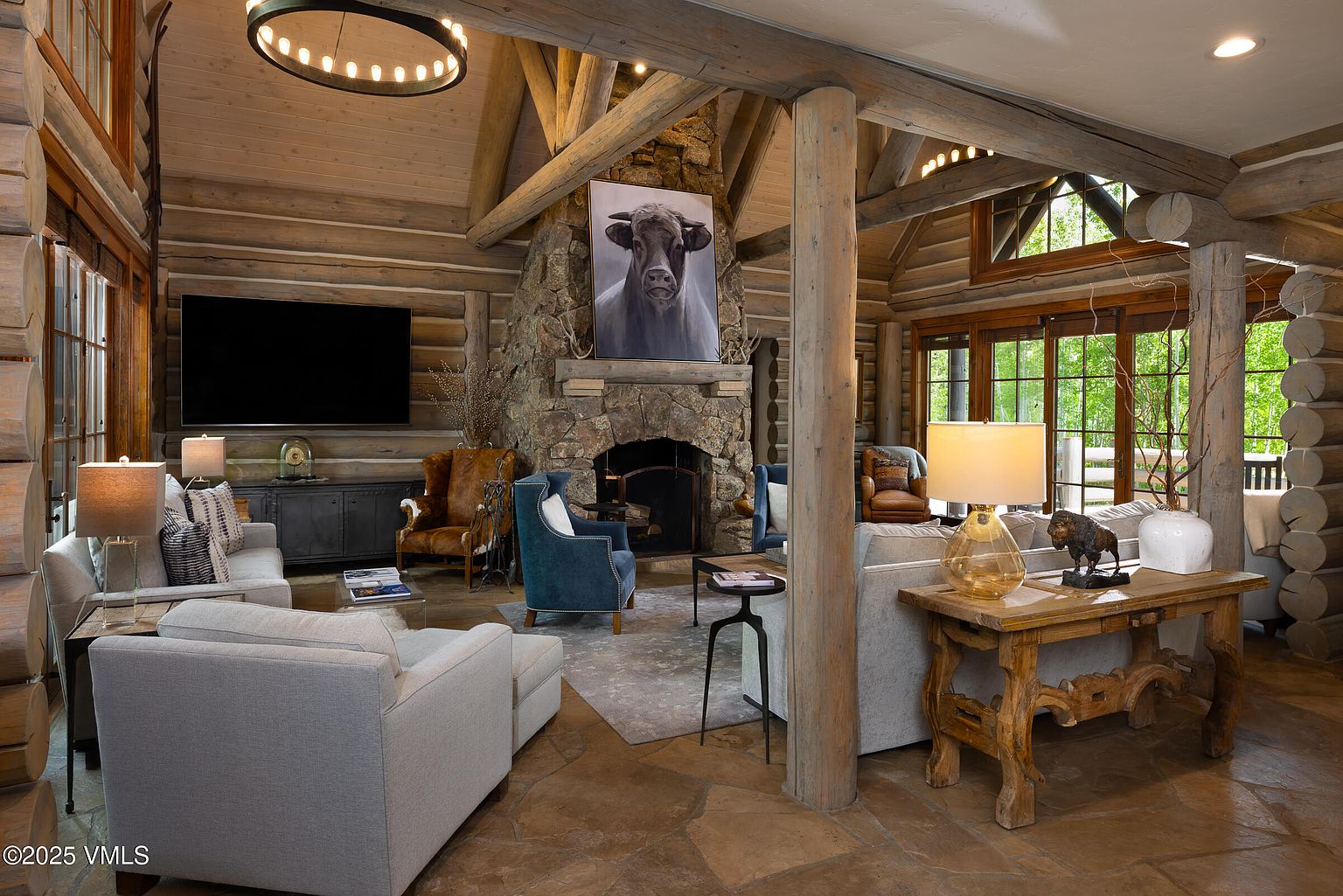 This is a spacious living room with a rustic, lodge-like aesthetic. The room features log walls, a stone fireplace with a large bull portrait above it, and comfortable seating arrangements including sofas and armchairs. A large chandelier hangs from the ceiling, and the room is filled with natural light from the windows, creating a warm and inviting atmosphere.