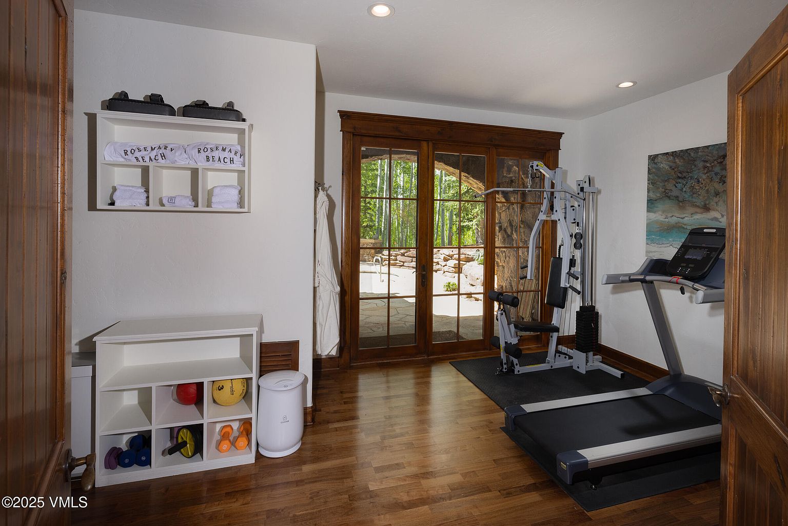This is an interior shot of a home gym featuring hardwood floors and white walls. The room includes a multi-station weight machine, a treadmill, and a storage unit with exercise equipment. Large glass doors provide natural light and a view of the outdoors, enhancing the workout environment.