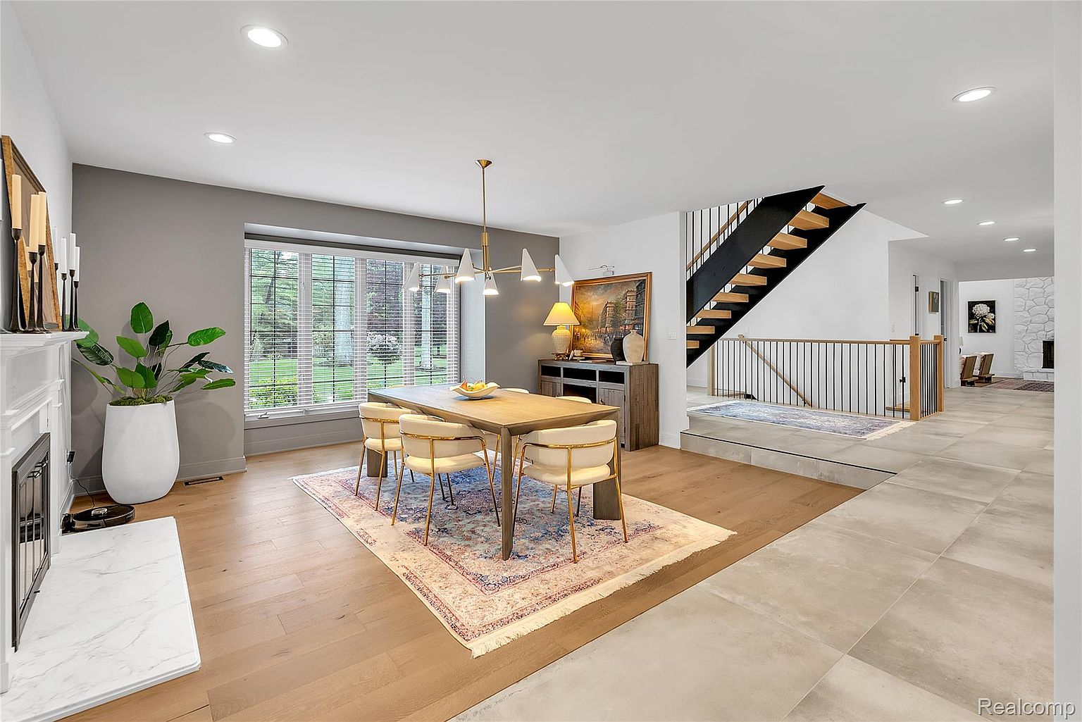This interior shot showcases a dining room with a modern aesthetic. A wooden dining table is surrounded by four chairs with gold frames and light-colored upholstery, set upon a patterned rug. The room features a fireplace, a large window with blinds, and a staircase in the background, blending contemporary design with comfortable living.