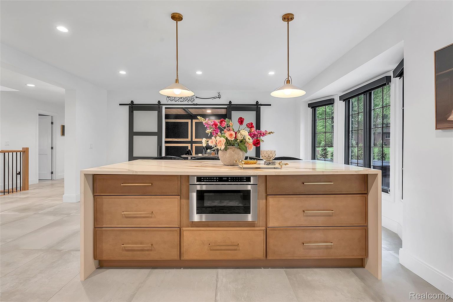 This interior shot showcases a modern kitchen island with light wood cabinetry, stainless steel microwave, and a light-colored countertop, possibly marble. Two pendant lights hang above the island, and a vase of colorful flowers adds a touch of elegance. The kitchen features a sliding barn door and large windows that provide natural light, enhancing the bright and airy feel of the space.