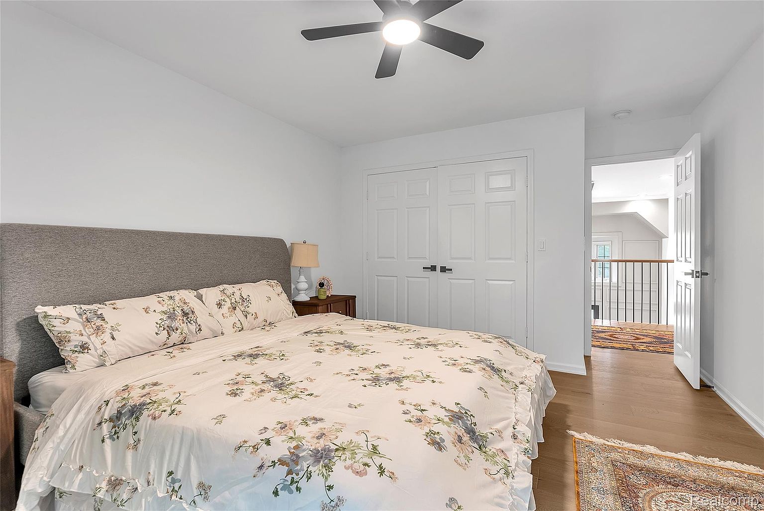 This is a bright and airy bedroom featuring a bed with a floral patterned comforter and a gray upholstered headboard. The room has white walls, a ceiling fan, and a double-door closet. A glimpse into the hallway reveals hardwood floors and a decorative rug, adding warmth to the space.