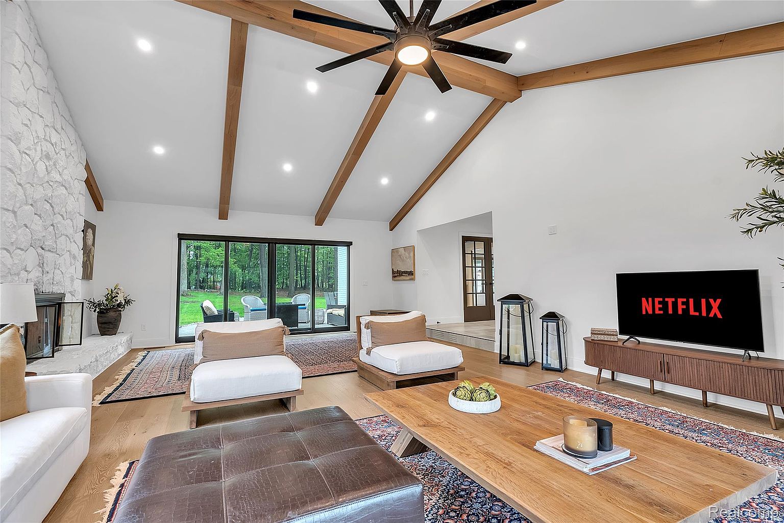 This is an interior shot of a living room featuring a high vaulted ceiling with wooden beams and recessed lighting. The room is decorated in a modern style with white walls, light wood flooring, and a stone fireplace. There is a large sliding glass door that leads to an outdoor area, and the furniture includes white armchairs, a leather ottoman, and a wooden coffee table. A television displaying the Netflix logo is visible against the wall.
