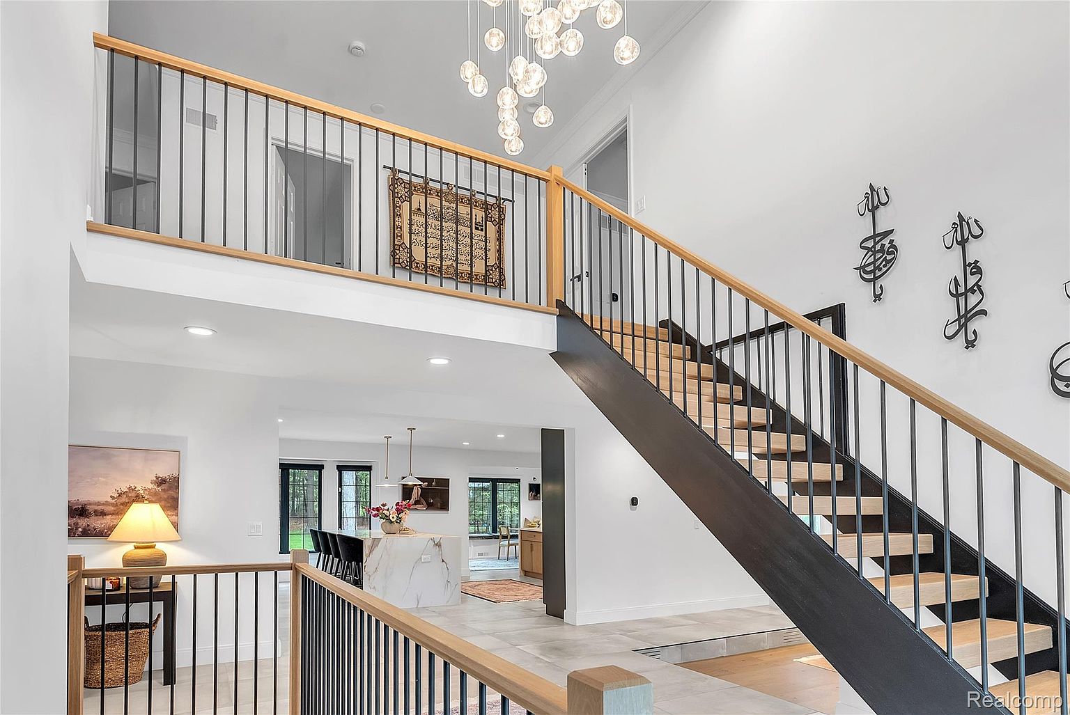 This interior shot showcases a modern home's grand foyer with a striking staircase featuring black metal railings and light wood treads. The open-concept design allows a glimpse into the kitchen area, highlighting a marble island and contemporary lighting. The space is bright and airy, emphasizing the home's sophisticated and welcoming atmosphere.