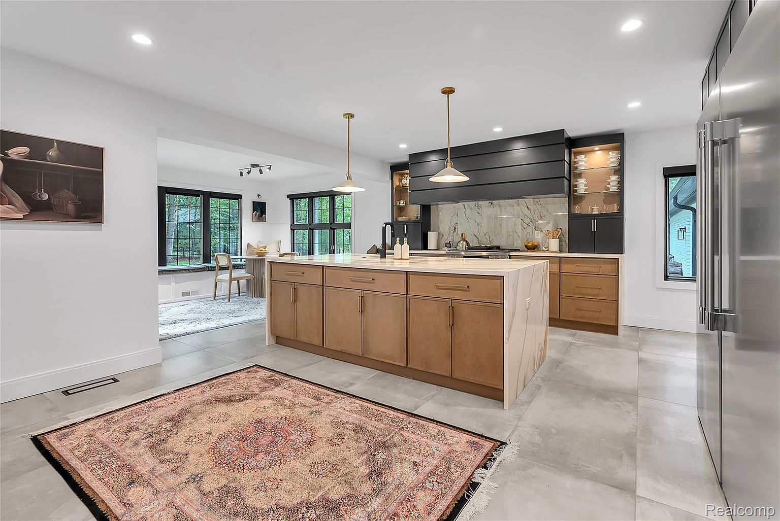 This is a bright and spacious kitchen featuring a large central island with a light countertop and wood-paneled cabinets. The kitchen also includes stainless steel appliances, a black range hood, and marble backsplash. The open layout connects to a dining area with large windows, creating a welcoming and modern space.