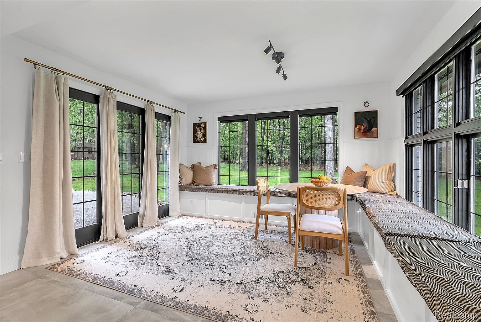 This bright and airy dining area features a built-in window seat with comfortable cushions, a round table with two chairs, and large windows offering views of the lush greenery outside. The room is decorated with neutral tones and a patterned rug, creating a cozy and inviting atmosphere. The black window frames and door frames add a modern touch to the space.