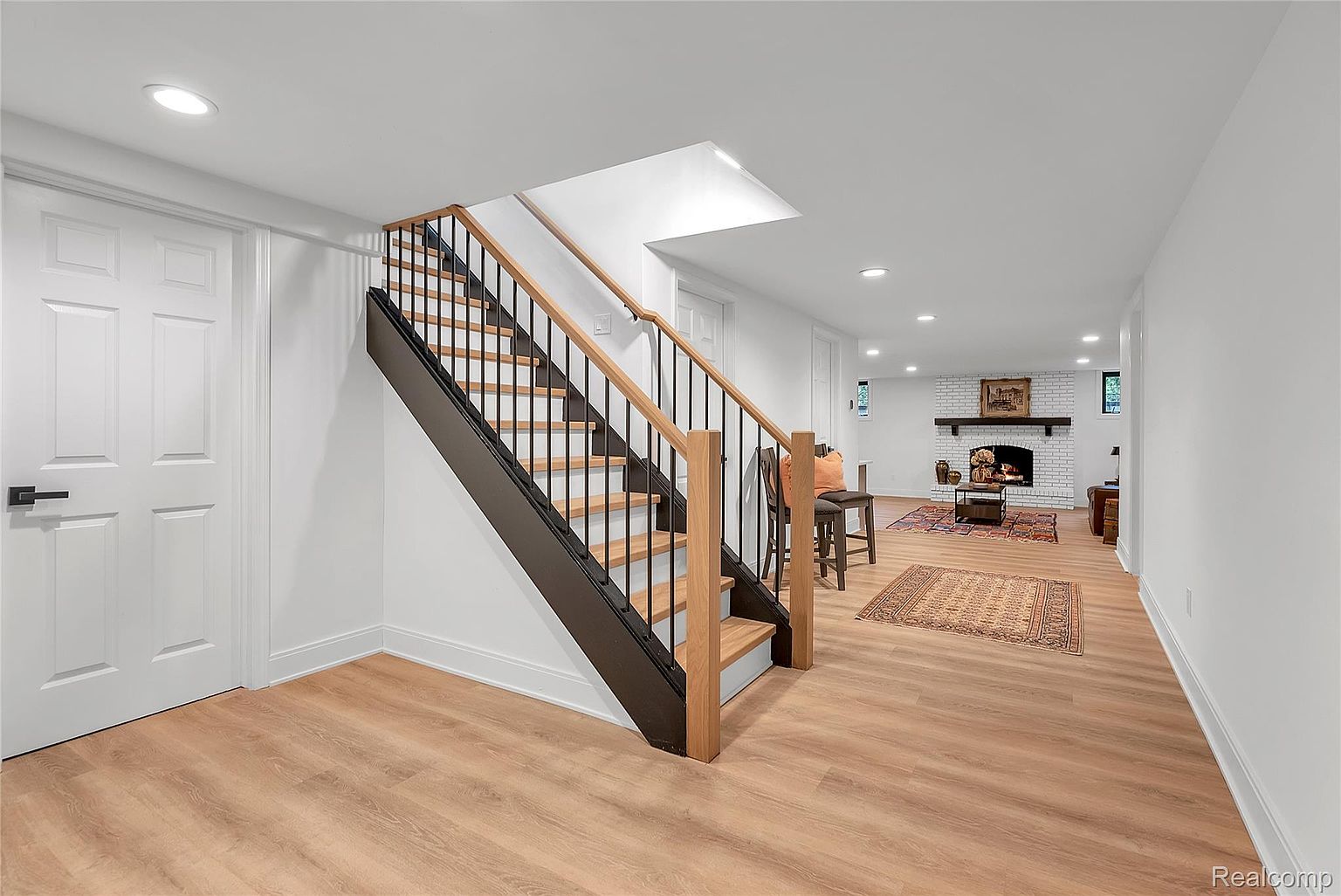 This is a well-lit basement featuring a staircase with wooden steps and black metal railings. The flooring is light-colored wood, and the walls are painted white. In the background, there is a fireplace with a white brick surround and a dark mantel, along with some seating and decorative items, creating a cozy and inviting atmosphere.
