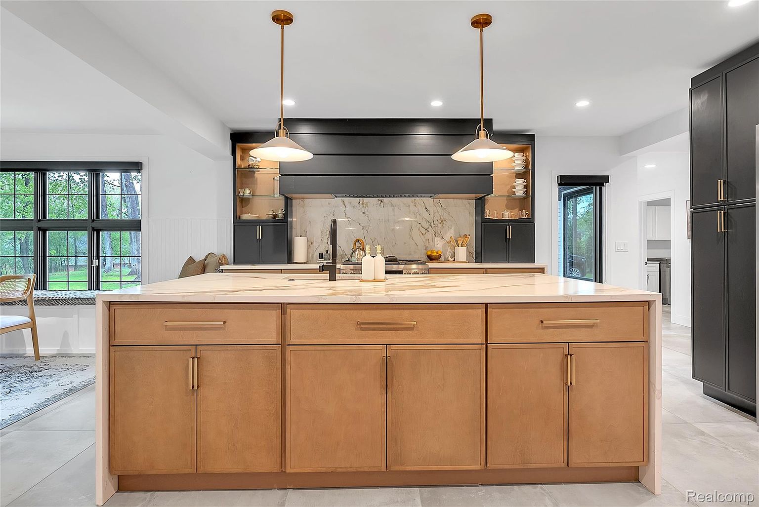 This is a bright and modern kitchen featuring a large island with light wood cabinetry and a marble countertop. The kitchen also includes dark gray cabinets, stainless steel appliances, and stylish pendant lighting. The space is well-lit and appears spacious, creating a welcoming atmosphere.