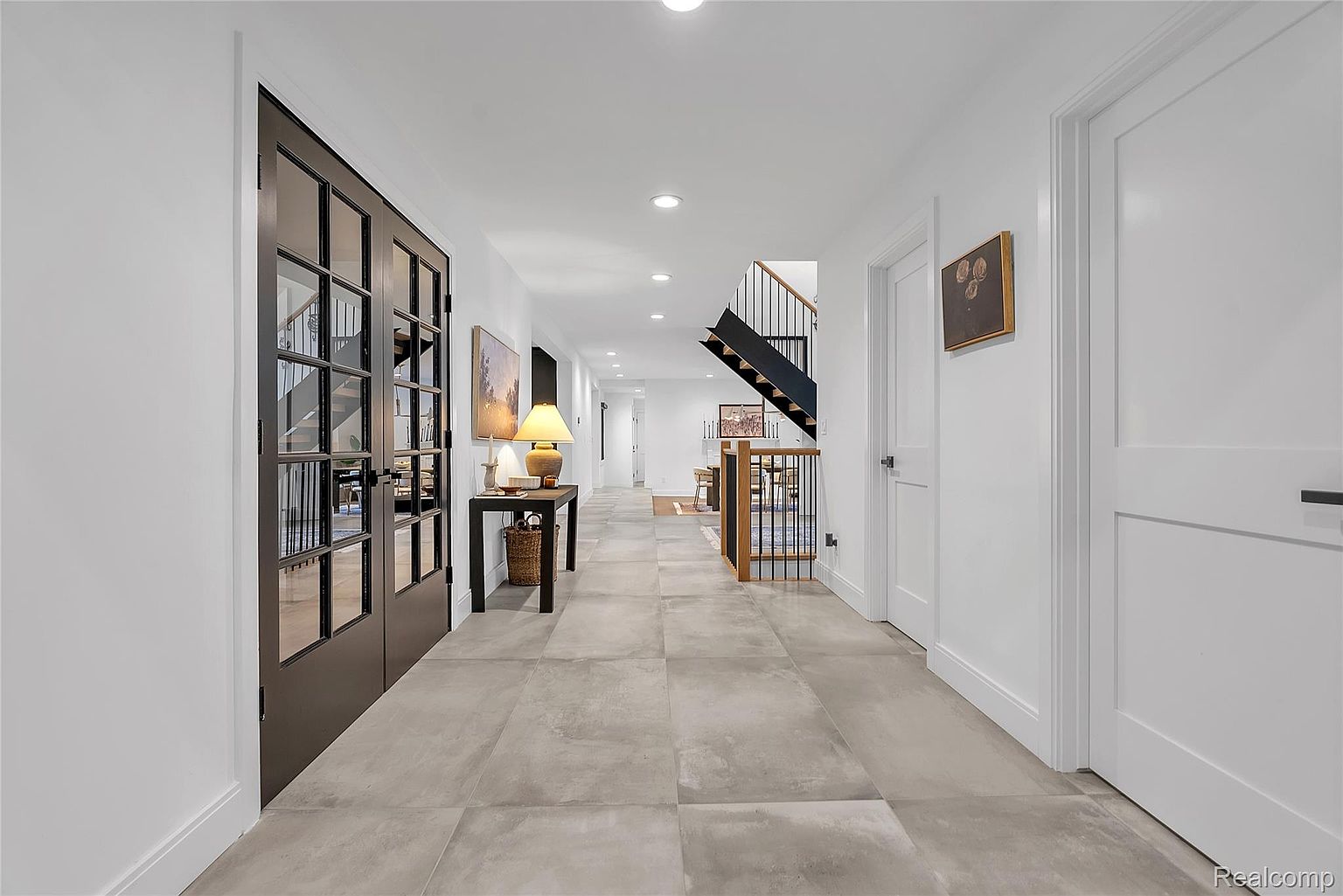 This interior shot showcases a long hallway with light gray tile flooring and white walls, creating a bright and spacious feel. A black-framed glass door is on the left, and a staircase with black metal railings and wood accents is visible further down the hall. The hallway is well-lit with recessed lighting, and artwork adorns the walls, adding a touch of elegance.