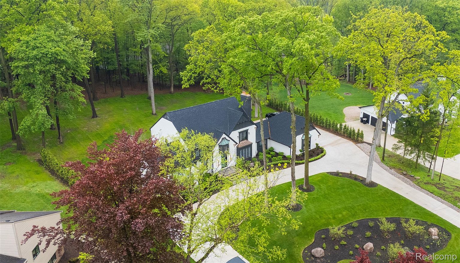 This aerial view showcases a stunning white house with a dark roof, surrounded by lush greenery and mature trees. A winding driveway leads to the house and a detached garage, highlighting the property's spaciousness and privacy. The well-manicured lawn and landscaping add to the overall curb appeal, creating an inviting and luxurious estate.