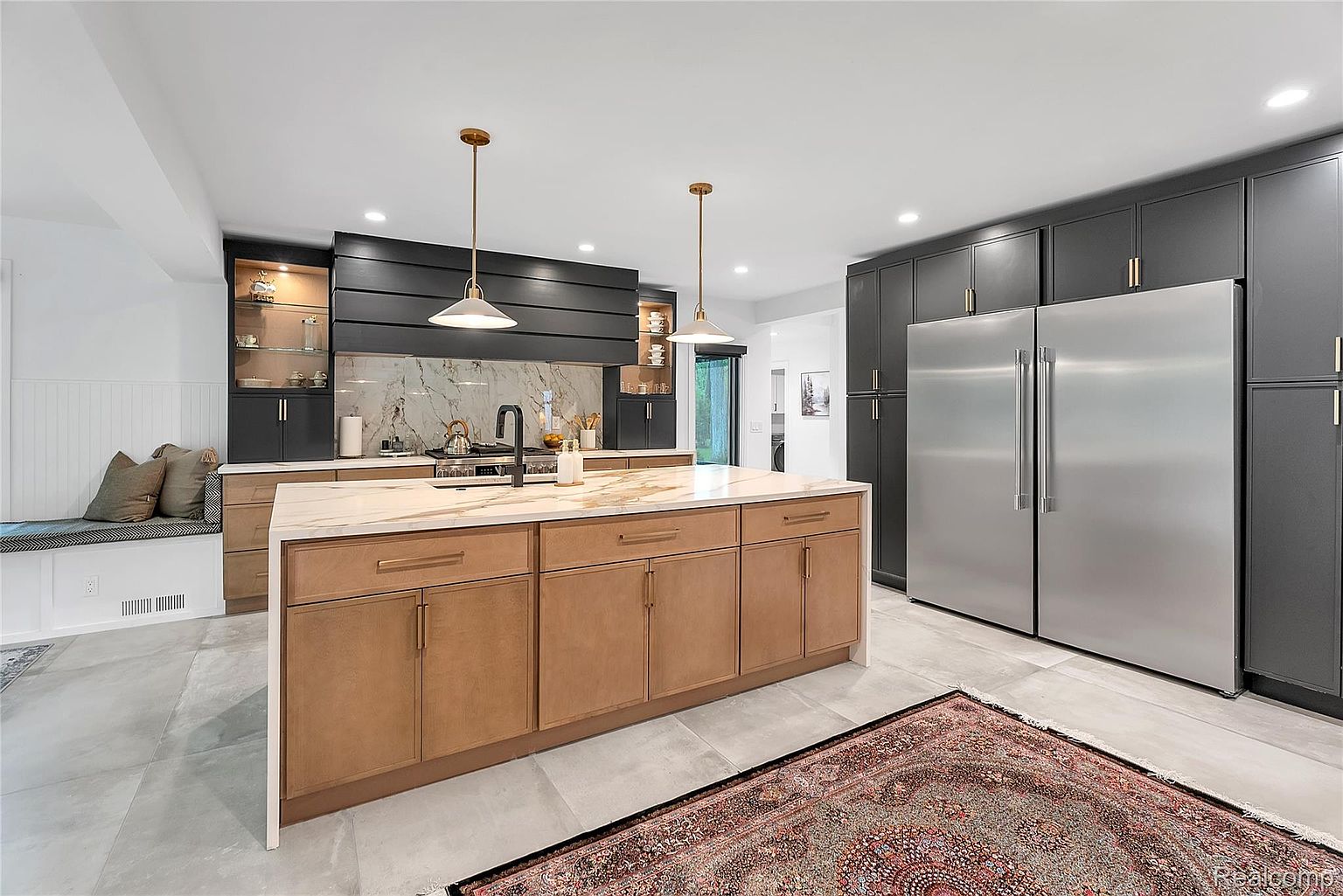 This is a modern kitchen featuring a large island with a light countertop and wood-toned cabinetry. The kitchen also includes dark gray cabinets, stainless steel appliances, and pendant lighting. A patterned rug adds warmth to the gray tile flooring, and a built-in bench provides a cozy seating area.