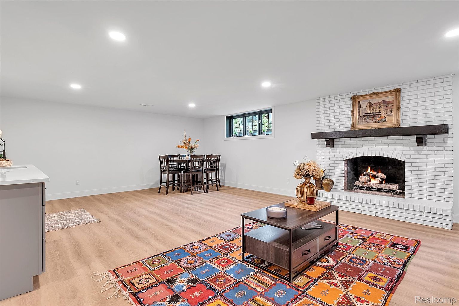 This is an interior shot of a living room featuring a white brick fireplace with a dark wood mantel and a framed painting above. A colorful patterned rug sits in front of the fireplace, with a dark wood coffee table placed on top. The room also includes a dining area with a dark wood table and chairs, and light wood flooring throughout.