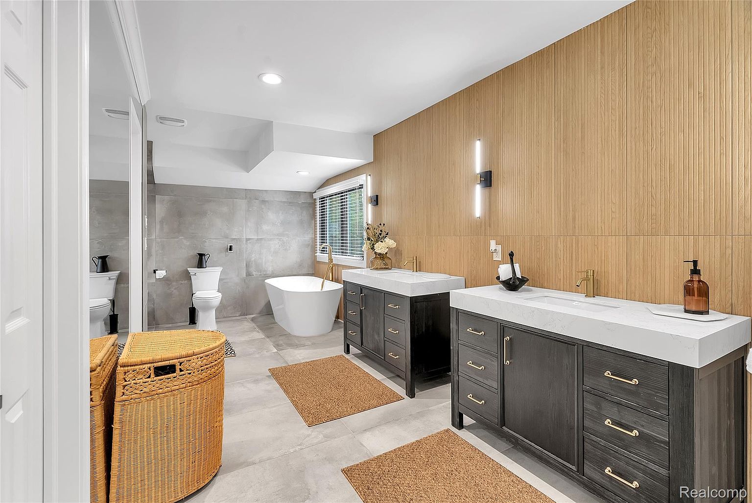 This is a primary bathroom featuring a modern design with a freestanding bathtub, dual vanities with white countertops and dark wood cabinetry, and a separate toilet area. The walls are a combination of wood paneling and large gray tiles, creating a luxurious and spa-like atmosphere. The perspective is from the doorway, showcasing the entire layout of the bathroom.