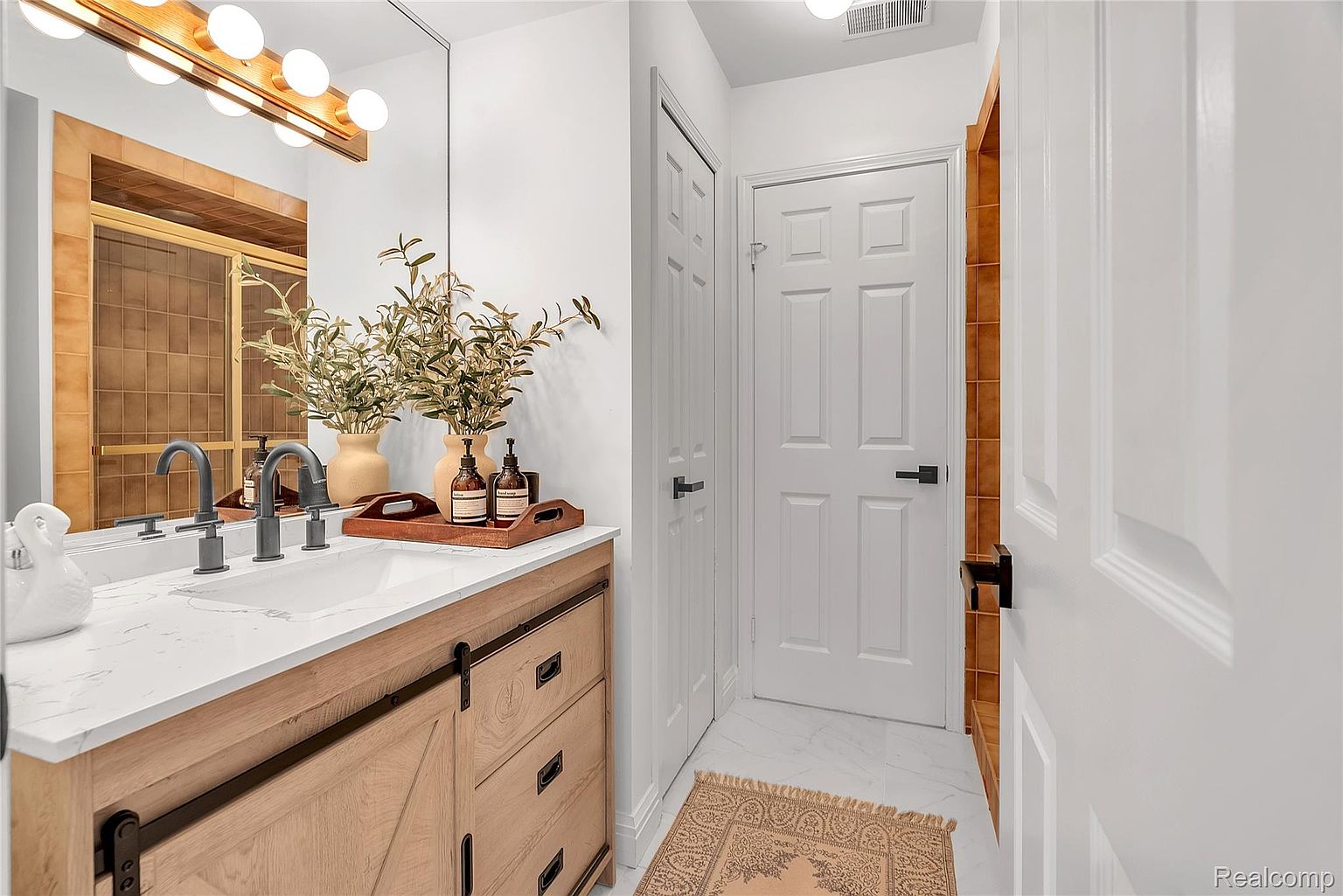 This is a well-lit bathroom featuring a modern vanity with a light wood finish and a white marble countertop. The vanity has a sliding barn door style cabinet and drawers with black hardware. A large mirror reflects the vanity and a tiled shower, while white walls and marble flooring contribute to a clean and contemporary aesthetic.