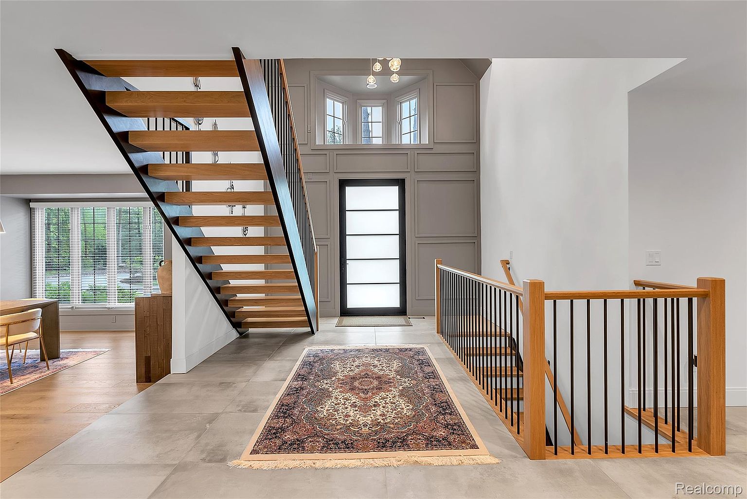 This interior shot showcases a modern entryway and staircase. The floating wooden stairs with black metal railings create a striking architectural feature. A patterned rug adds warmth to the tiled floor, while the front door with frosted glass panels allows natural light to filter in, creating a bright and inviting space.