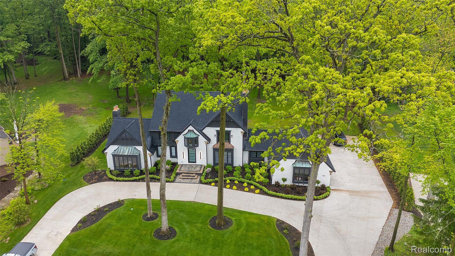 This aerial view showcases a stunning two-story home with a dark roof and white exterior, complemented by black trim and accents. The property features a well-manicured lawn, mature trees, and a winding driveway leading to the house. The overall impression is one of luxury and privacy, set within a lush, green landscape.