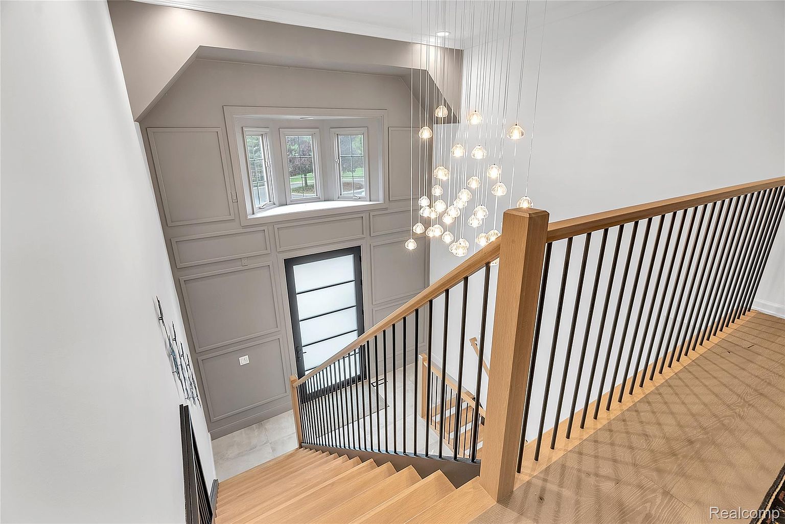 This interior shot showcases a modern staircase with wooden steps and black metal railings, leading down from an upper level. A striking chandelier hangs above, adding a touch of elegance to the space. The walls are painted in neutral tones, and a window above the front door allows natural light to flood the area, creating a bright and inviting atmosphere.
