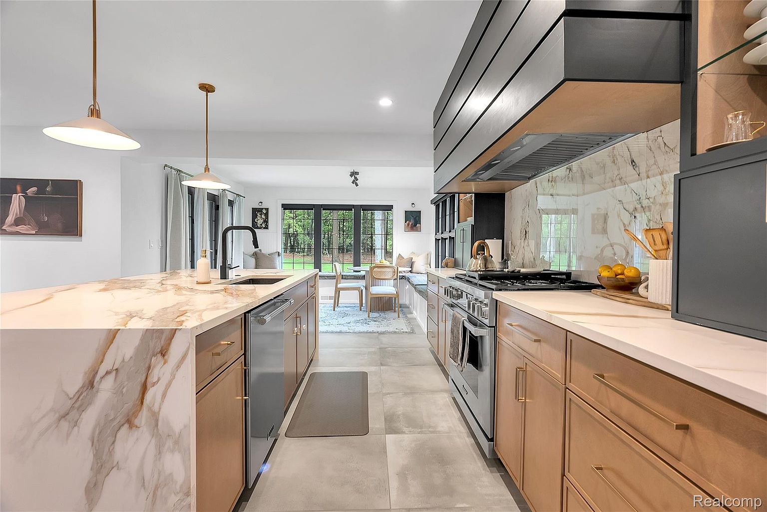 This is a bright and modern kitchen featuring a large island with a marble countertop and a sink. The kitchen has wooden cabinets with gold hardware, stainless steel appliances, and a marble backsplash. The kitchen opens up to a dining area with a view of the backyard, creating an open and inviting space.