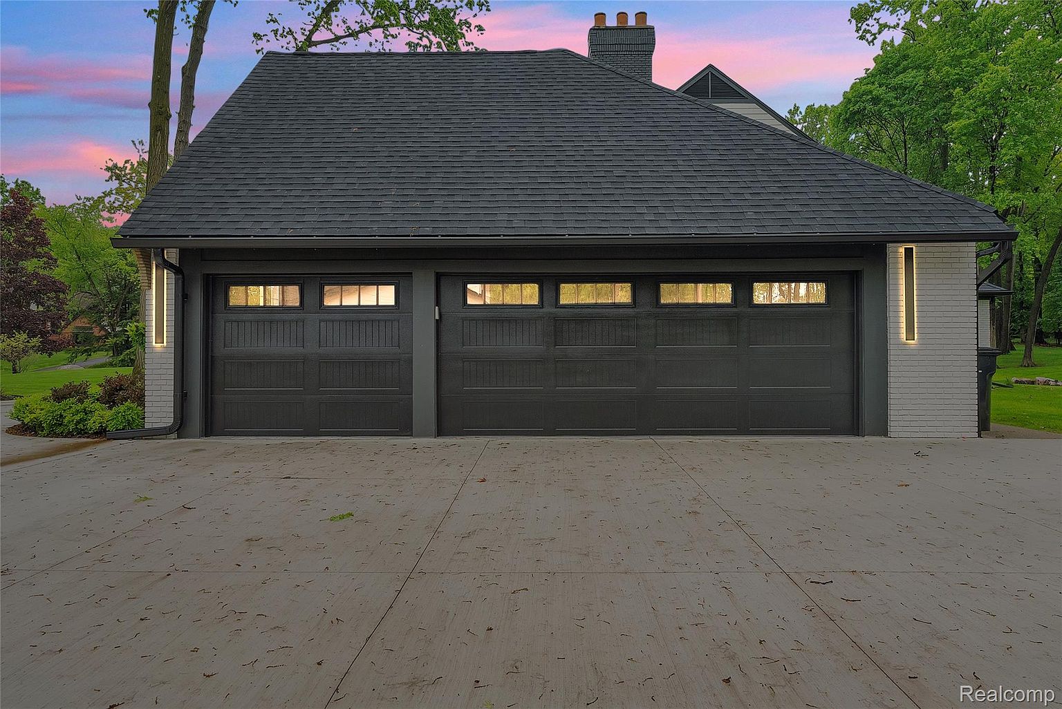 This image showcases a modern garage exterior with two dark-colored garage doors, each featuring small windows near the top. The garage is attached to a house with a dark roof and light-colored brick accents on the sides. The driveway is concrete and appears clean, and the overall impression is one of contemporary design and functionality.