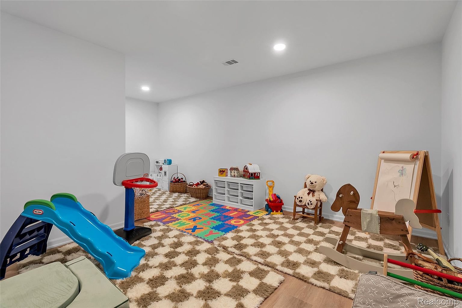 This is a well-lit basement playroom featuring a variety of children's toys, including a slide, basketball hoop, and rocking horse. The room is carpeted with a patterned rug and has white walls, creating a bright and inviting space. The overall impression is a clean and organized area perfect for family activities.