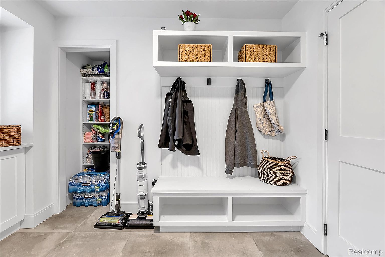 This is an interior shot of a well-organized laundry room or mudroom. The room features built-in shelving with woven baskets, a coat rack with hanging jackets and a tote bag, and a bench with storage underneath. A closet with shelving is visible to the left, and the flooring appears to be tile, creating a clean and functional space.