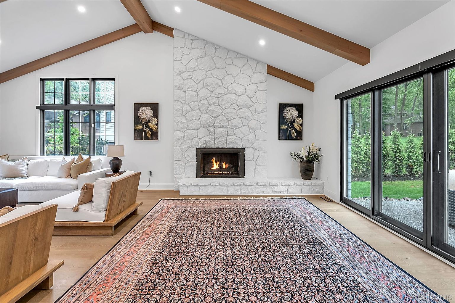 This is a bright and airy living room featuring a stone fireplace as a focal point, complemented by wooden ceiling beams and hardwood floors. The room is furnished with a comfortable white sofa and a stylish wooden chaise lounge, creating a welcoming and modern atmosphere. Large sliding glass doors provide ample natural light and a view of the outdoors.