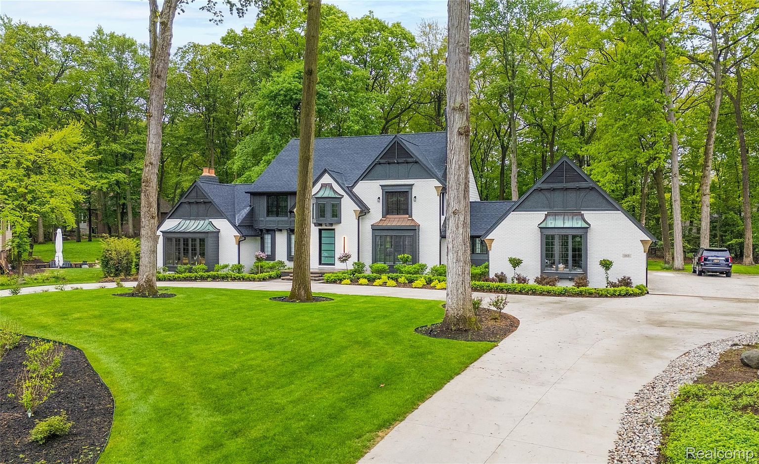 This is a front exterior view of a stately two-story home with a well-manicured lawn and mature trees. The house features a white brick facade with contrasting dark gray trim and a dark roof. A curved driveway leads to the front of the house, and landscaping includes neatly trimmed bushes and flower beds, creating a welcoming and luxurious curb appeal.