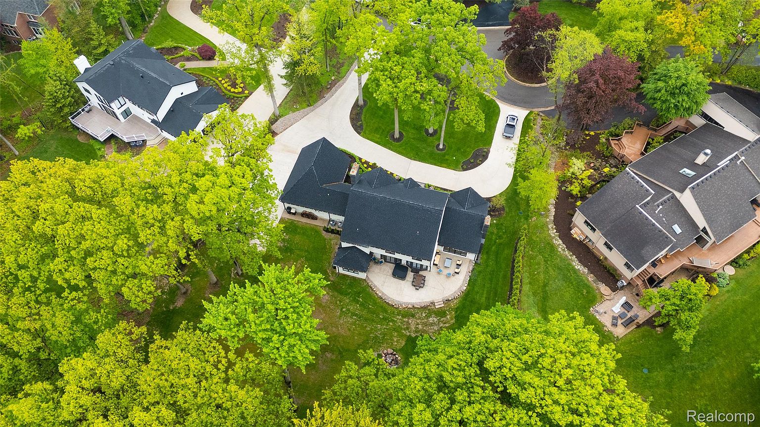 This aerial shot showcases a luxurious home nestled among lush greenery. The property features a dark roof, a circular patio with outdoor furniture, and a winding driveway leading to the house. The surrounding landscape includes mature trees and well-maintained lawns, creating a serene and private setting.