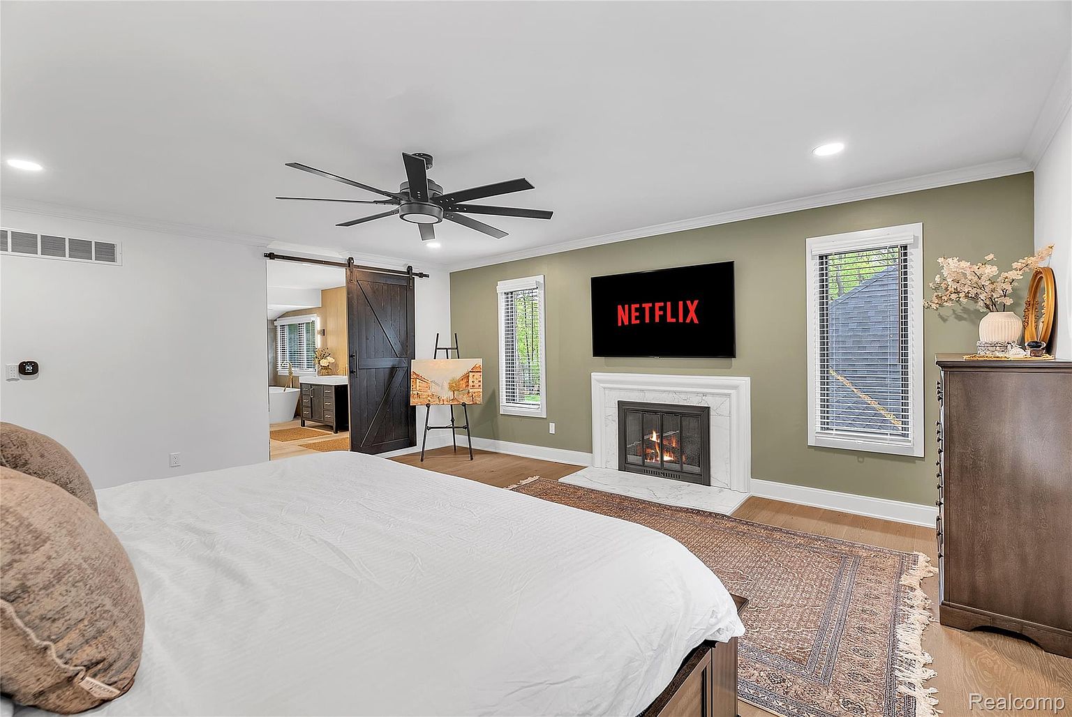 This is a primary bedroom featuring a king-size bed with white linens, a dark wood dresser, and a fireplace with a television mounted above it. The room has a modern aesthetic with a sliding barn door leading to the bathroom and an easel with a painting. The color palette is neutral with a pop of green on the wall behind the fireplace.