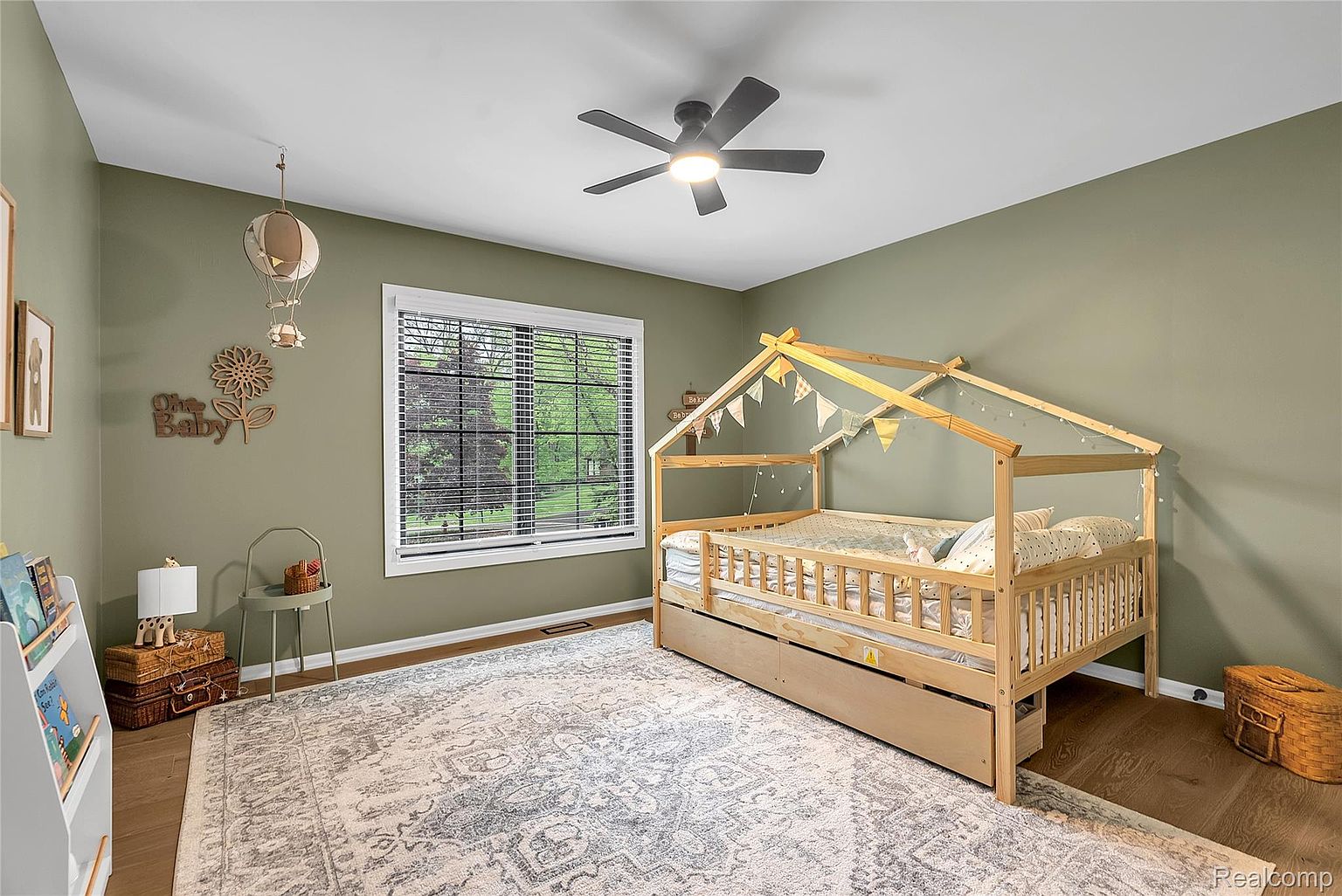 This is a charming bedroom designed for a child, featuring a unique house-frame bed adorned with decorative lights and pennants. The room is painted in a muted green tone, complemented by a patterned area rug and hardwood flooring. Natural light streams in through a window, creating a warm and inviting atmosphere.