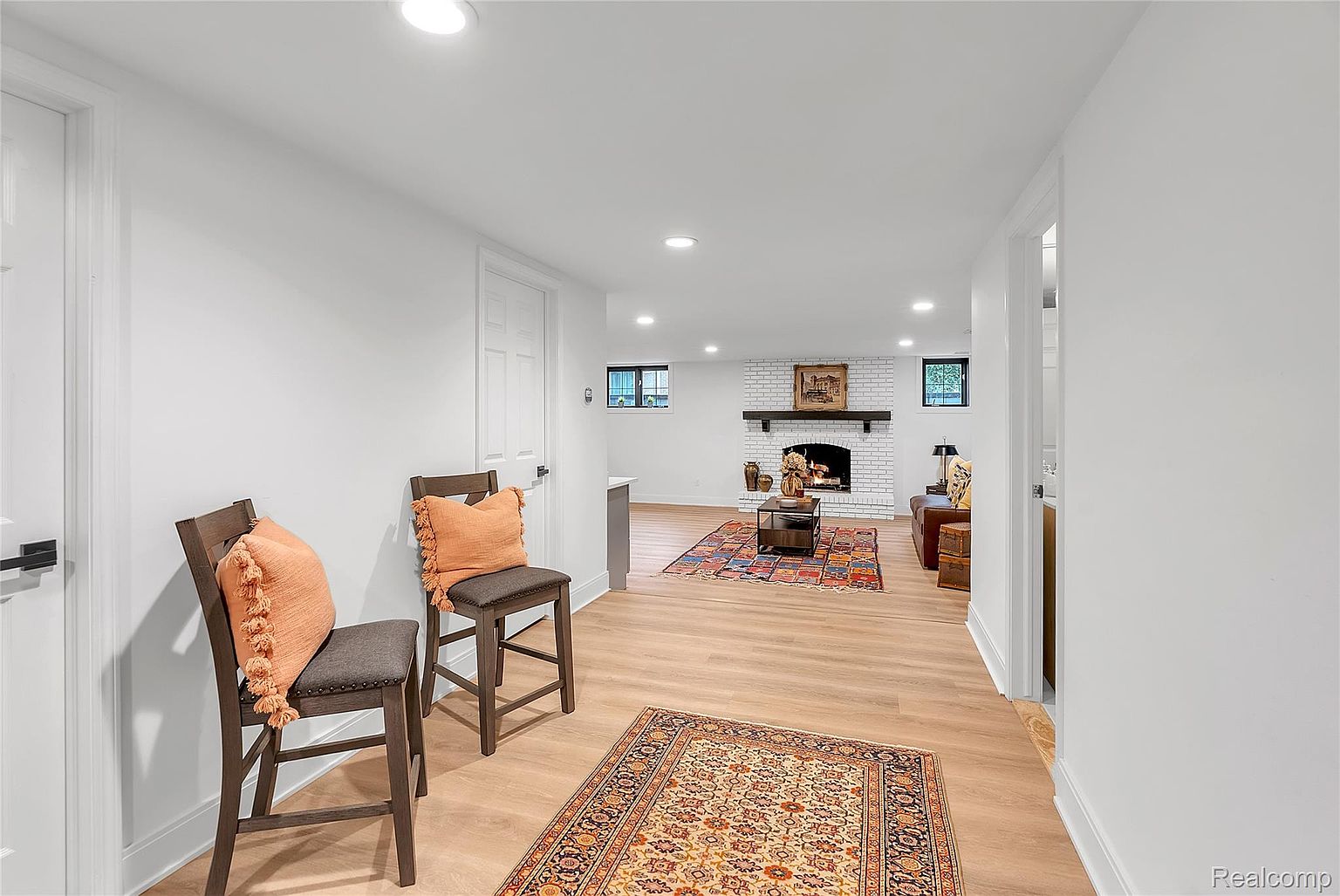 This interior shot showcases a cozy living room with a white brick fireplace and dark wood mantle. The room features hardwood floors, a patterned area rug, and comfortable seating. Two decorative chairs with orange pillows are in the foreground, adding a pop of color and warmth to the space.