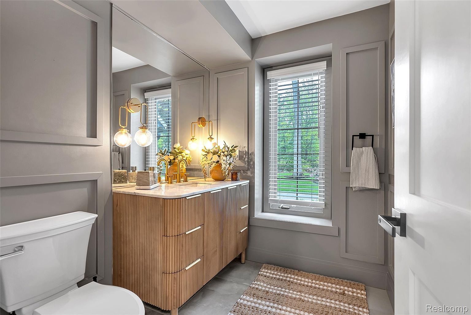 This is a well-lit bathroom featuring a modern vanity with a light wood finish and a marble countertop. The walls are painted in a neutral gray tone, complemented by white trim and a matching rug. A window with blinds allows natural light to filter into the space, creating a bright and inviting atmosphere.