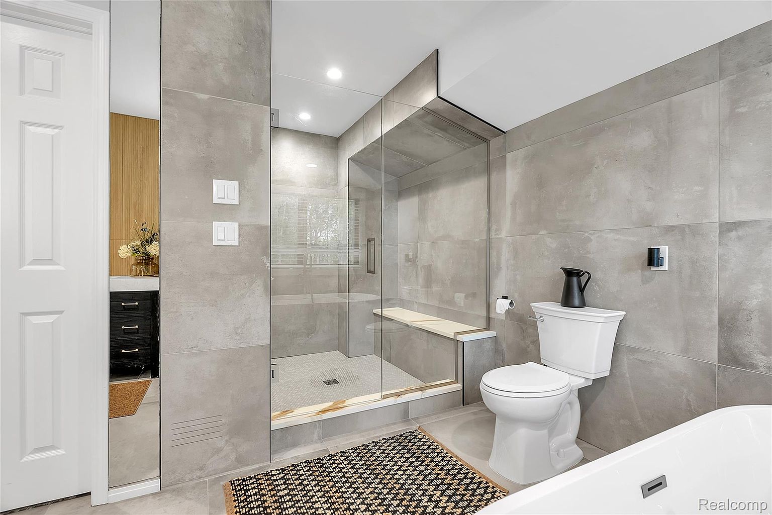 This is a modern primary bathroom featuring gray tile walls and flooring. A glass-enclosed shower with a built-in bench is visible, along with a toilet and a partial view of a bathtub. The space is well-lit and has a clean, contemporary aesthetic.