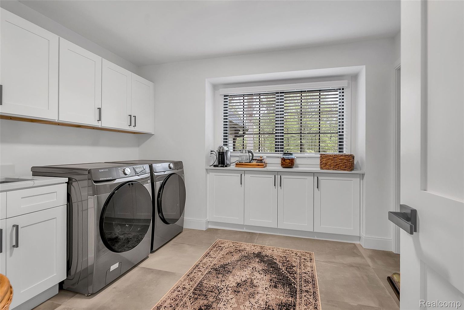 This is a well-organized laundry room featuring white cabinetry above and below a countertop, providing ample storage space. A modern, dark-colored Samsung washer and dryer set are prominently displayed, and a decorative rug adds a touch of warmth to the tiled floor. The room is brightly lit by natural light from a window with blinds.