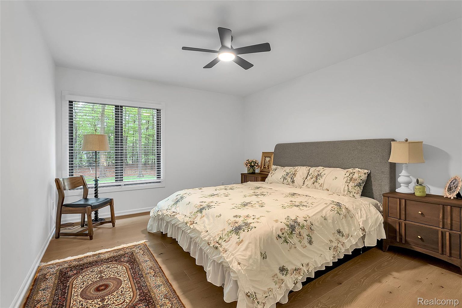 This is a well-lit primary bedroom featuring a queen-sized bed with a floral patterned comforter and a gray upholstered headboard. The room has hardwood floors, a ceiling fan, and a large window with blinds, providing natural light. Two wooden nightstands with lamps flank the bed, and a decorative rug adds warmth to the space.