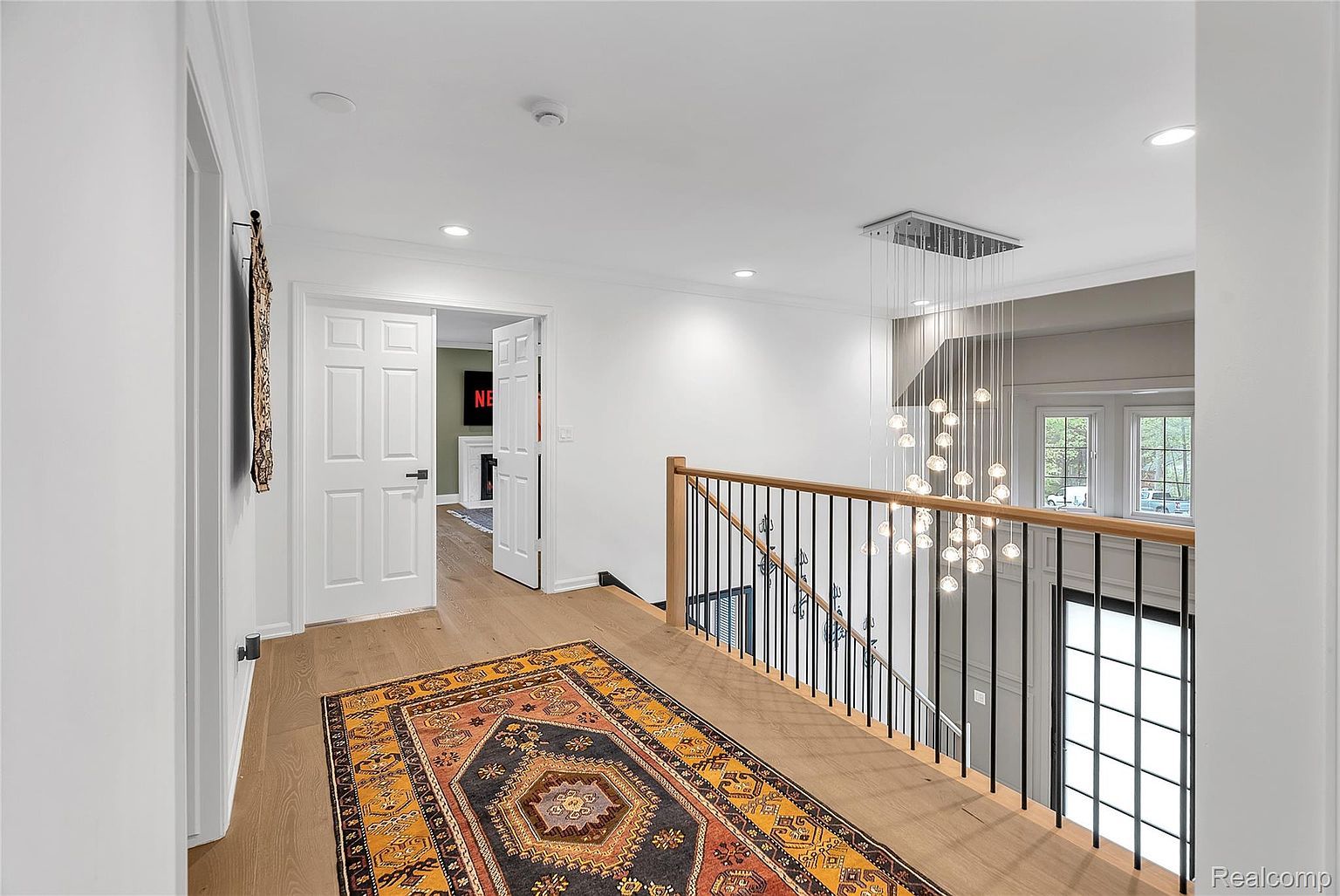 This interior shot showcases a bright hallway with a staircase. The hallway features light wood flooring, white walls, and a colorful patterned rug. A modern chandelier hangs above the staircase, which has black metal railings and a light wood handrail. The open doorway reveals a glimpse of a room with a television.