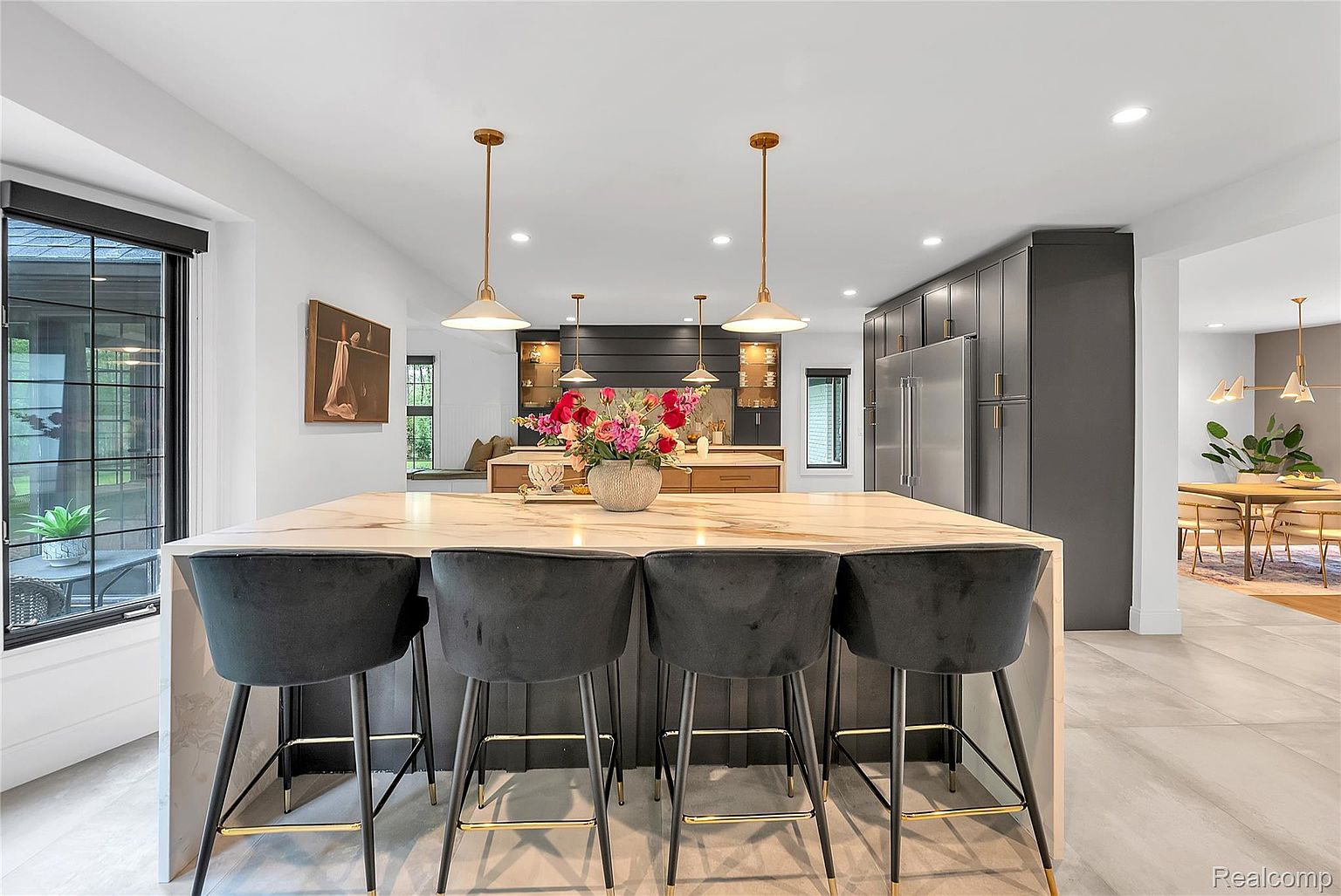 This is a well-lit, modern kitchen featuring a large island with a marble countertop and seating for four. The kitchen includes dark gray cabinetry, stainless steel appliances, and pendant lighting. A dining area is visible in the background, creating an open and inviting space.