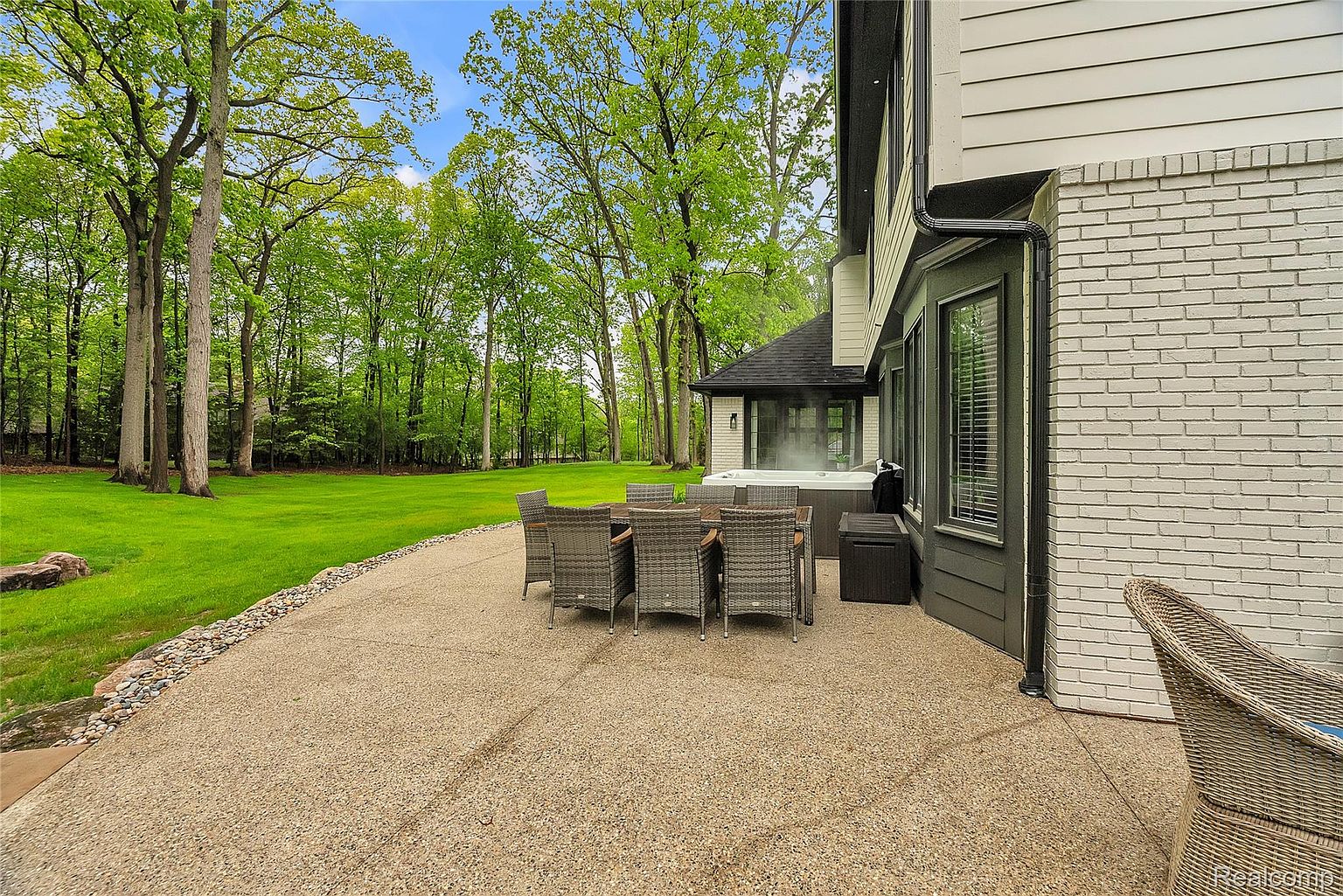 This image showcases a well-appointed patio area, perfect for outdoor entertaining. A dining set with seating for eight is centrally located, adjacent to a hot tub and a storage unit. The patio is constructed of exposed aggregate concrete and is bordered by a lush green lawn and mature trees, creating a private and serene backyard setting.