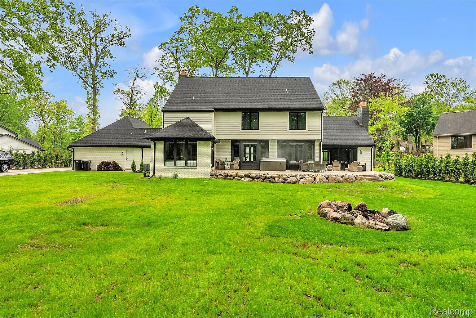 This is a rear view of a modern two-story home with a dark roof and light-colored siding. The backyard features a well-maintained green lawn, a stone-lined patio with outdoor furniture and a hot tub, and a fire pit area with large rocks. The landscaping includes mature trees and shrubs, creating a private and inviting outdoor space.
