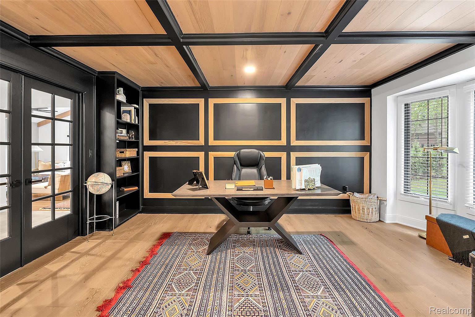 This is a well-appointed home office featuring dark walls with framed paneling, a light wood plank ceiling with dark beams, and hardwood floors. A large desk with a leather chair sits atop an ornate rug, and a built-in bookshelf adds storage. The room is illuminated by natural light from a window and a central ceiling light, creating a sophisticated and functional workspace.