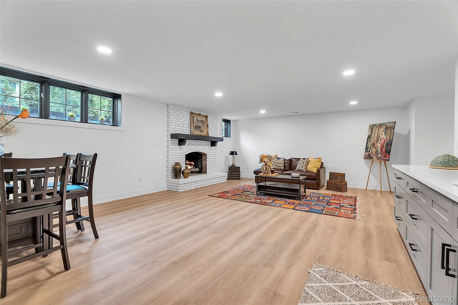 This is an interior shot of a living room featuring light wood flooring and white walls. A white brick fireplace with a dark mantle is a focal point, complemented by a brown leather sofa and a colorful area rug. An easel with a painting adds an artistic touch, and the room is well-lit with recessed lighting.