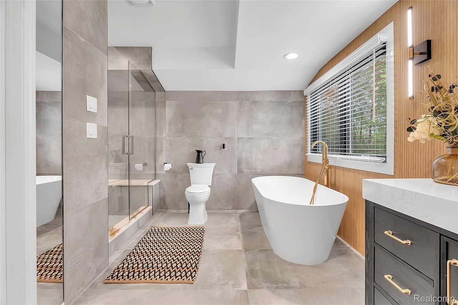 This is a modern primary bathroom featuring large gray tiles on the walls and floor, a freestanding white bathtub with a gold faucet, and a glass-enclosed shower. The room also includes a toilet, a dark vanity with a white countertop, and a window with blinds, creating a spa-like atmosphere. The perspective is from the doorway, showcasing the layout and high-end finishes.