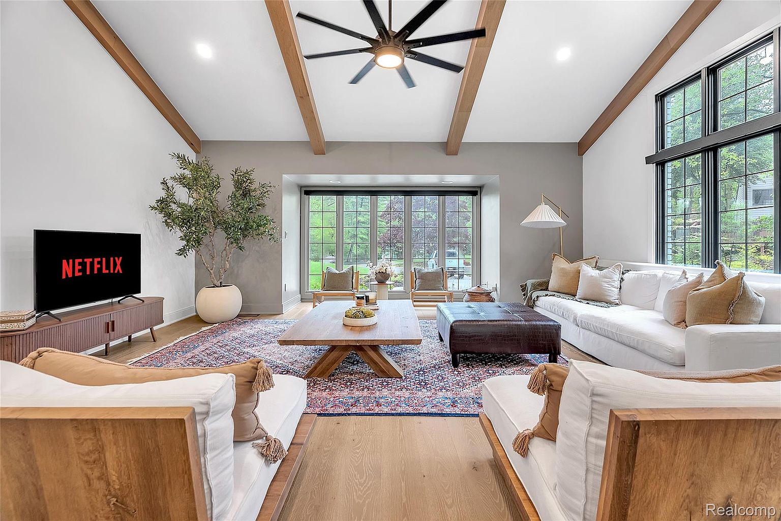This is a bright and spacious living room featuring a large window with a view of the outdoors. The room is decorated with a mix of modern and rustic elements, including wooden beams, a large area rug, and comfortable seating. A television is visible on a console, and a large plant adds a touch of nature to the space.