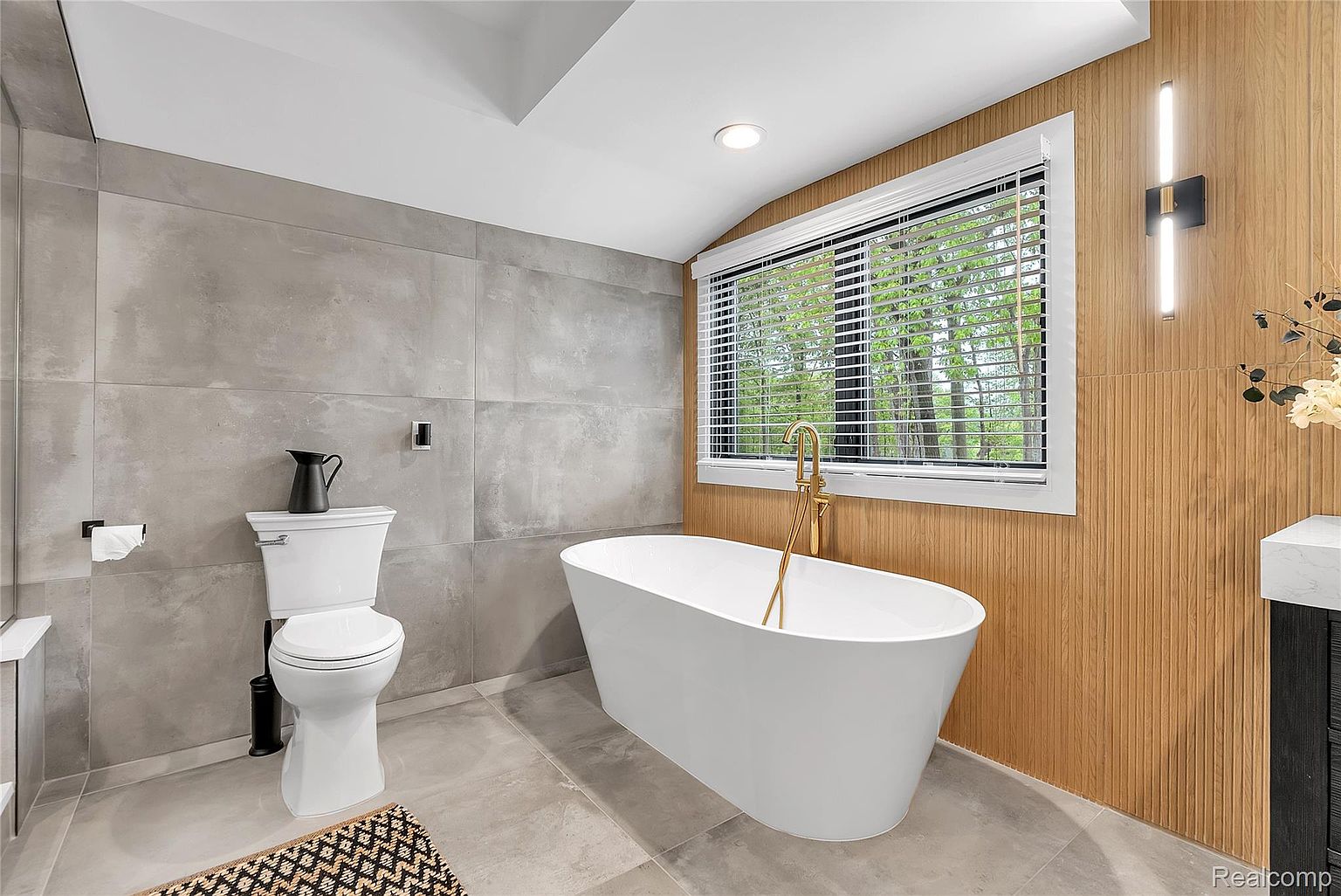 This is a modern primary bathroom featuring a freestanding white bathtub with a gold faucet, a toilet with a black pitcher on top, and a window with blinds. The walls are a mix of gray tile and wood paneling, complemented by gray tile flooring. The overall impression is clean, luxurious, and well-lit.
