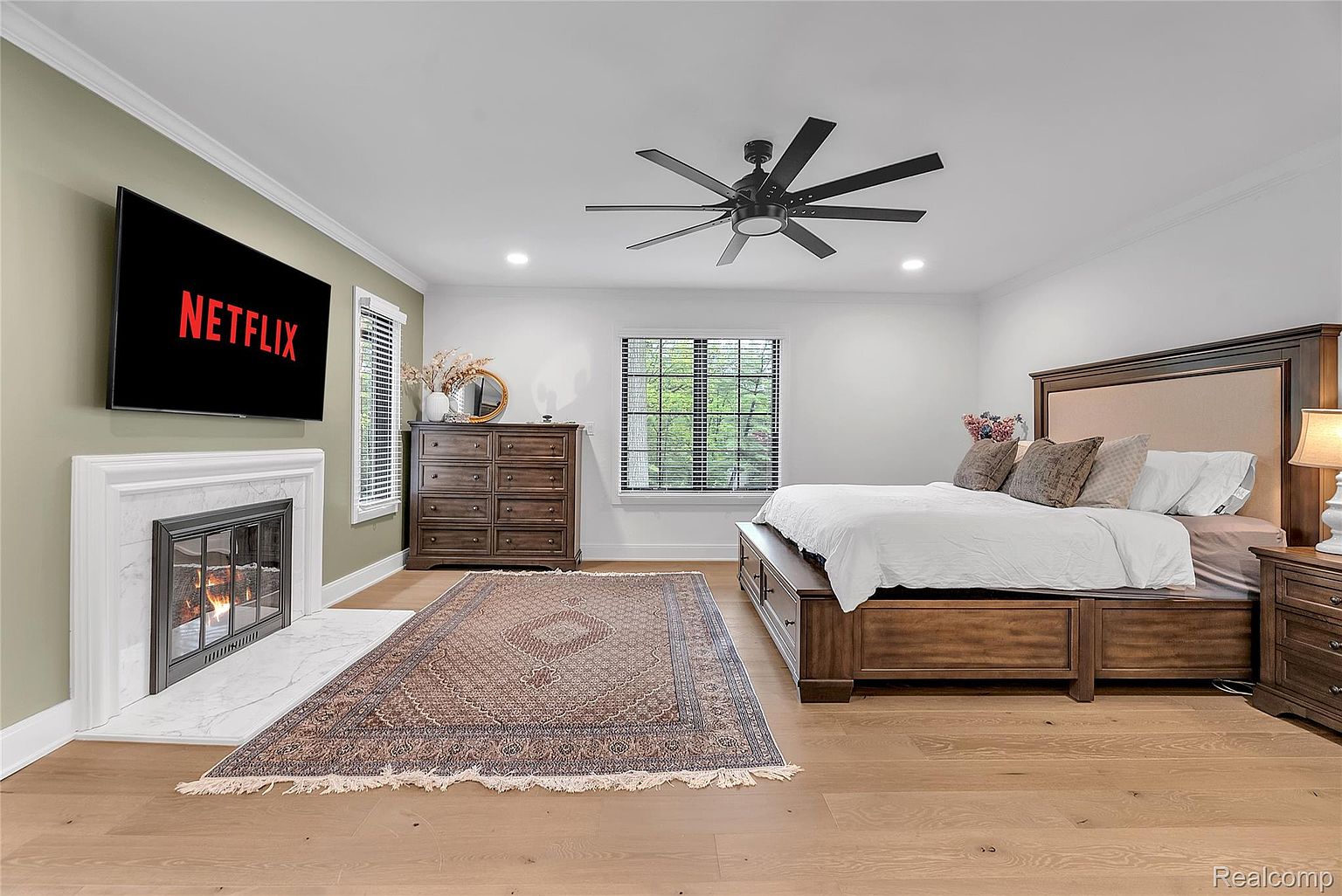 This is a primary bedroom featuring a large bed with a wooden frame and upholstered headboard, complemented by matching nightstands. A fireplace with a TV above it adds a cozy focal point, while a patterned rug anchors the space. The room is well-lit with natural light from a window and features a ceiling fan.