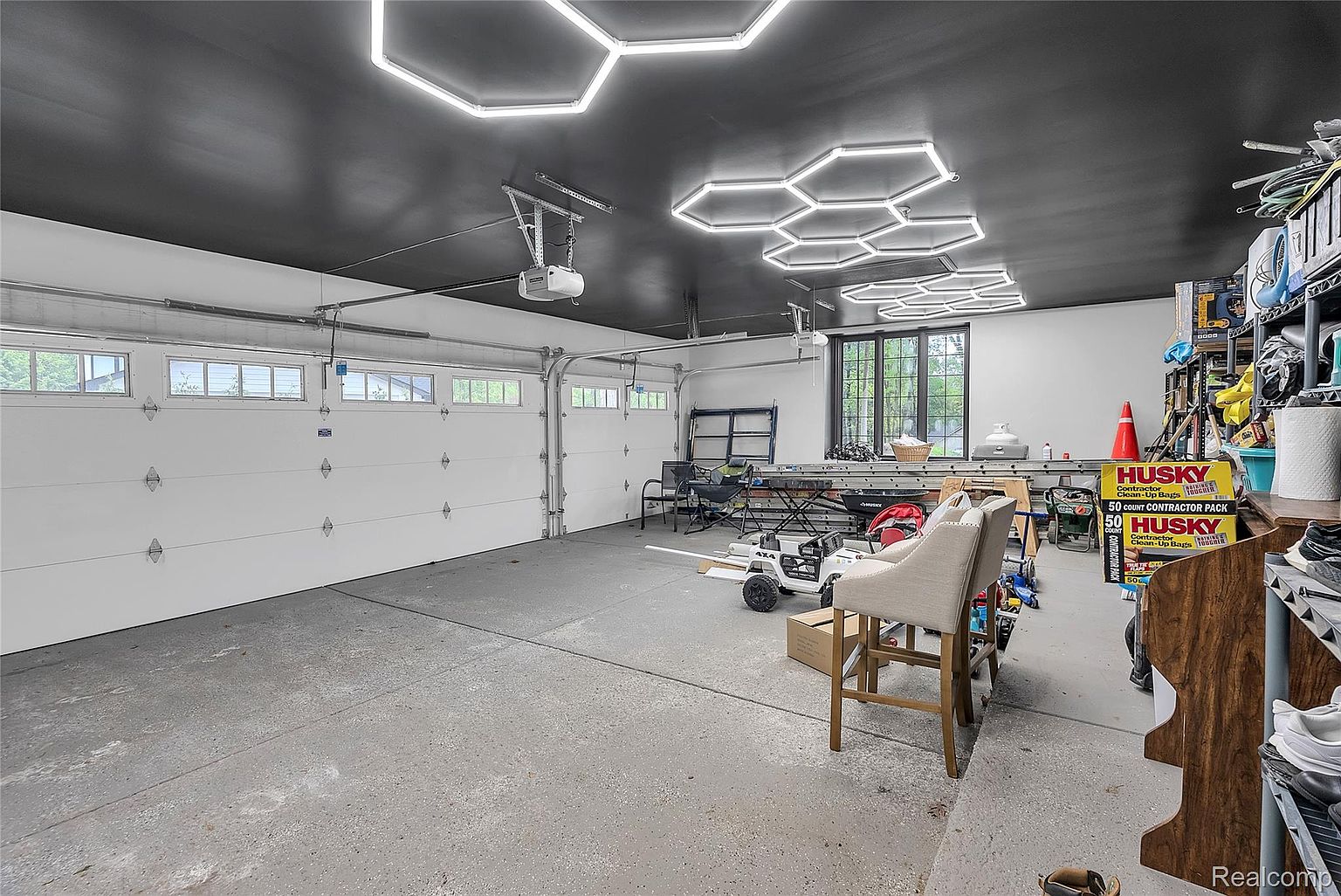 This image showcases a well-organized and modern garage interior. The garage features a black ceiling with unique hexagonal LED lighting fixtures, providing ample illumination. White garage doors with windows add natural light, and the gray epoxy flooring contributes to a clean and contemporary aesthetic.