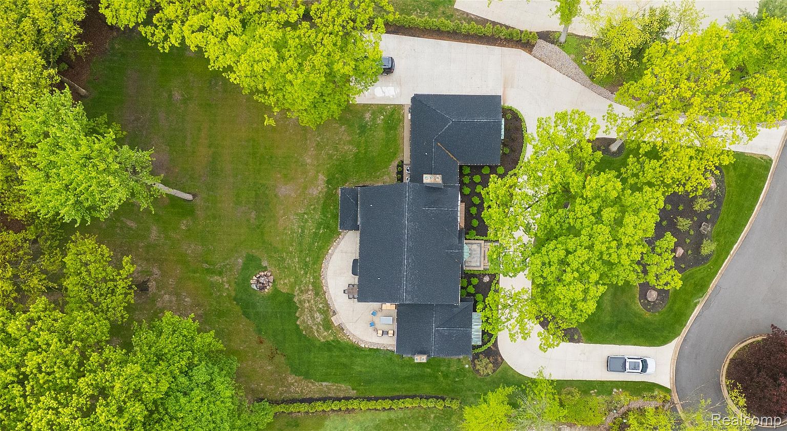This aerial shot showcases a well-manicured property featuring a large house with a dark roof, surrounded by lush green lawns and mature trees. A winding driveway leads to the house, and a patio area with outdoor furniture is visible. The overall impression is one of privacy and luxury.