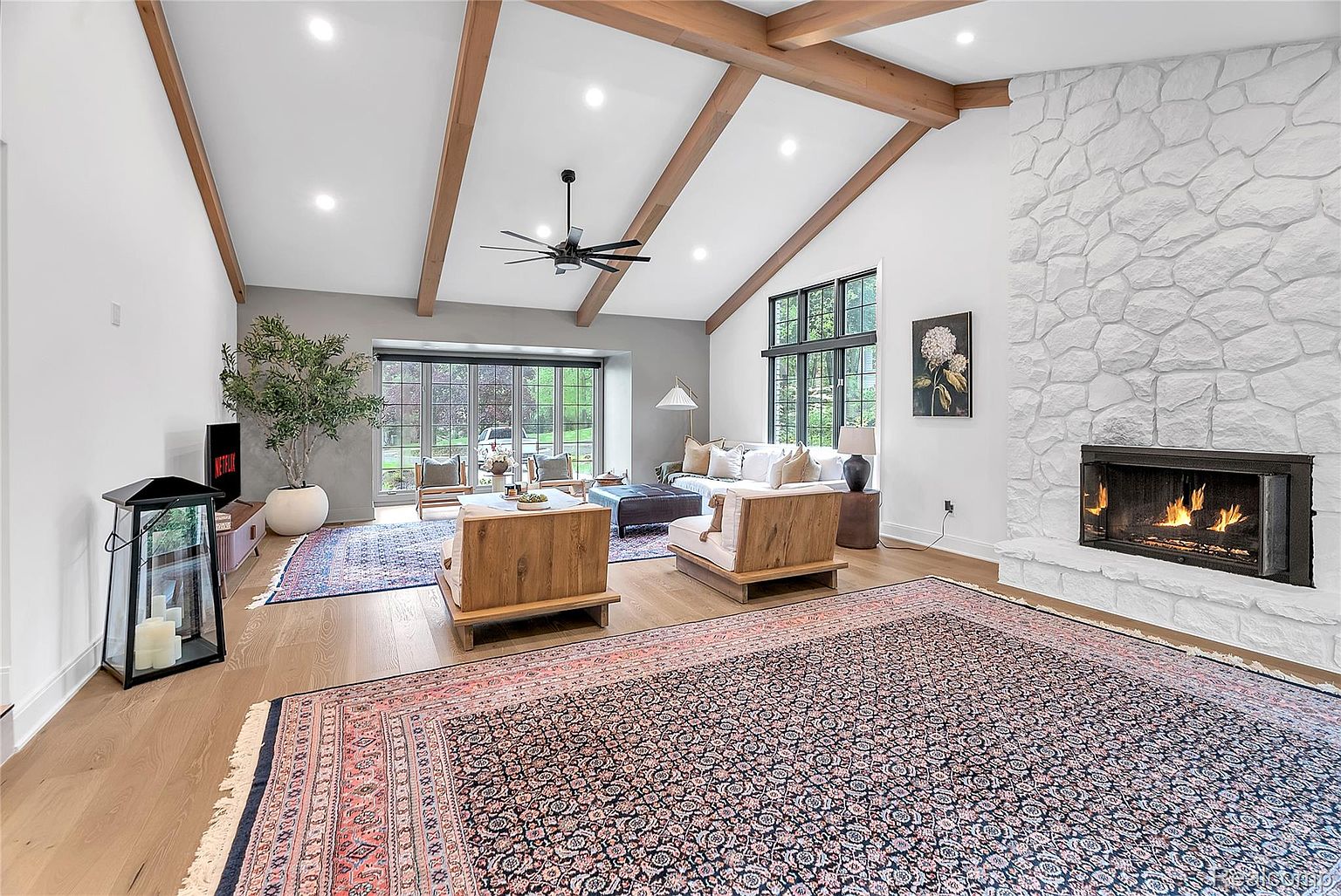 This is an interior shot of a living room featuring a high vaulted ceiling with wooden beams, a stone fireplace, and large windows that provide ample natural light. The room is furnished with modern wooden chairs, a sofa, and a large area rug, creating a warm and inviting atmosphere. The perspective is from the front of the room, showcasing the layout and design elements.
