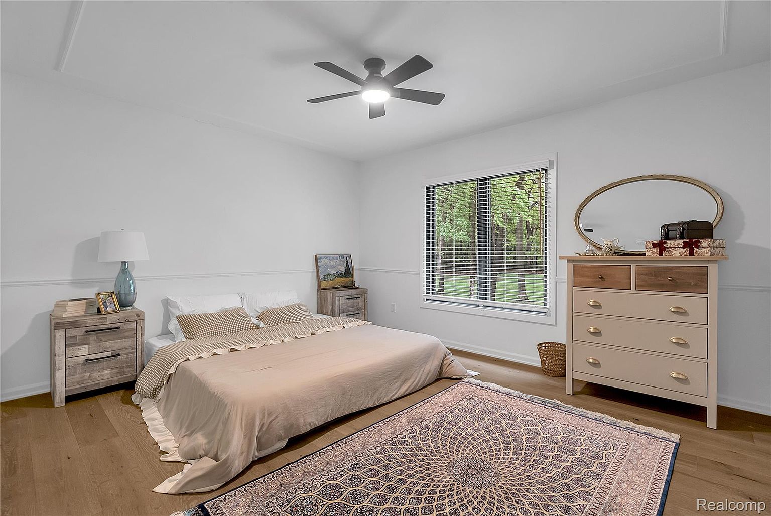This is a bright and airy primary bedroom featuring a neutral color palette. The room includes a bed with beige linens, a wooden nightstand with a lamp, and a dresser with a mirror. A large window provides natural light, and a patterned rug adds a touch of warmth to the hardwood floors.